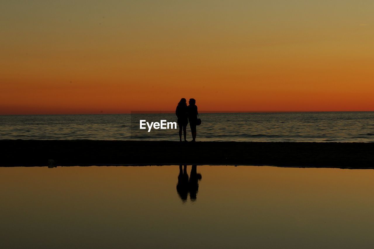 Silhouette women standing on beach against orange sky