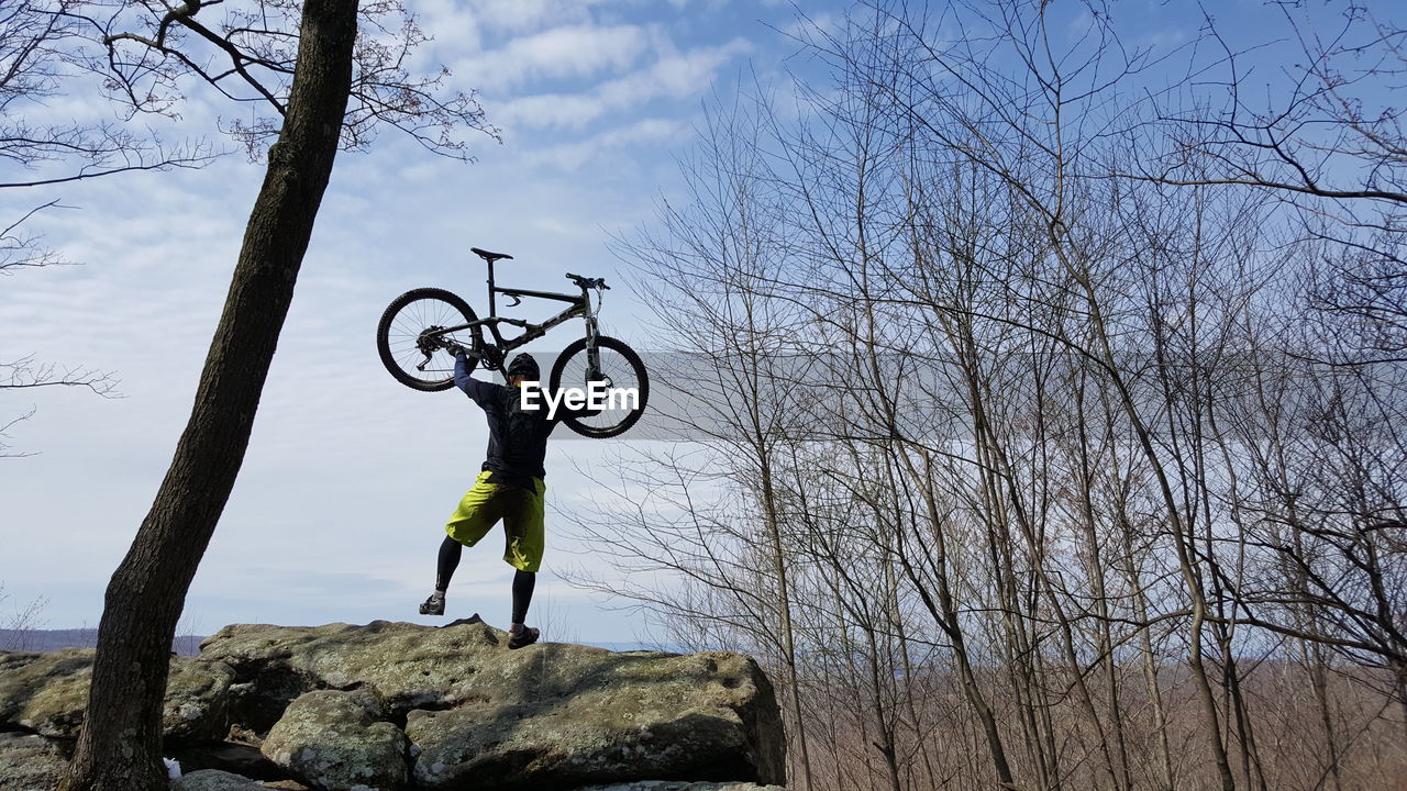 Rear view of man holding bicycle while standing on rock against sky