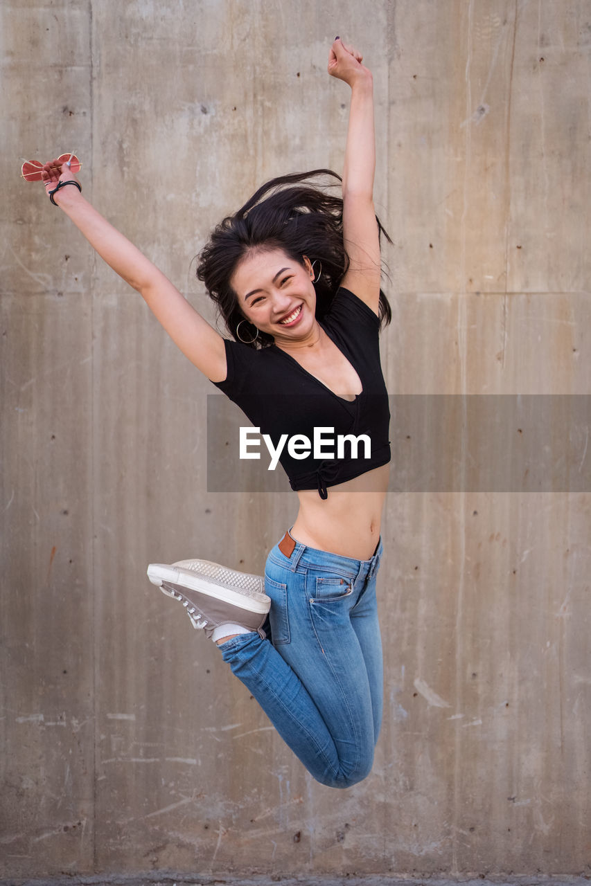 Side view of delighted asian female celebrating victory and jumping with raised arms while looking at camera