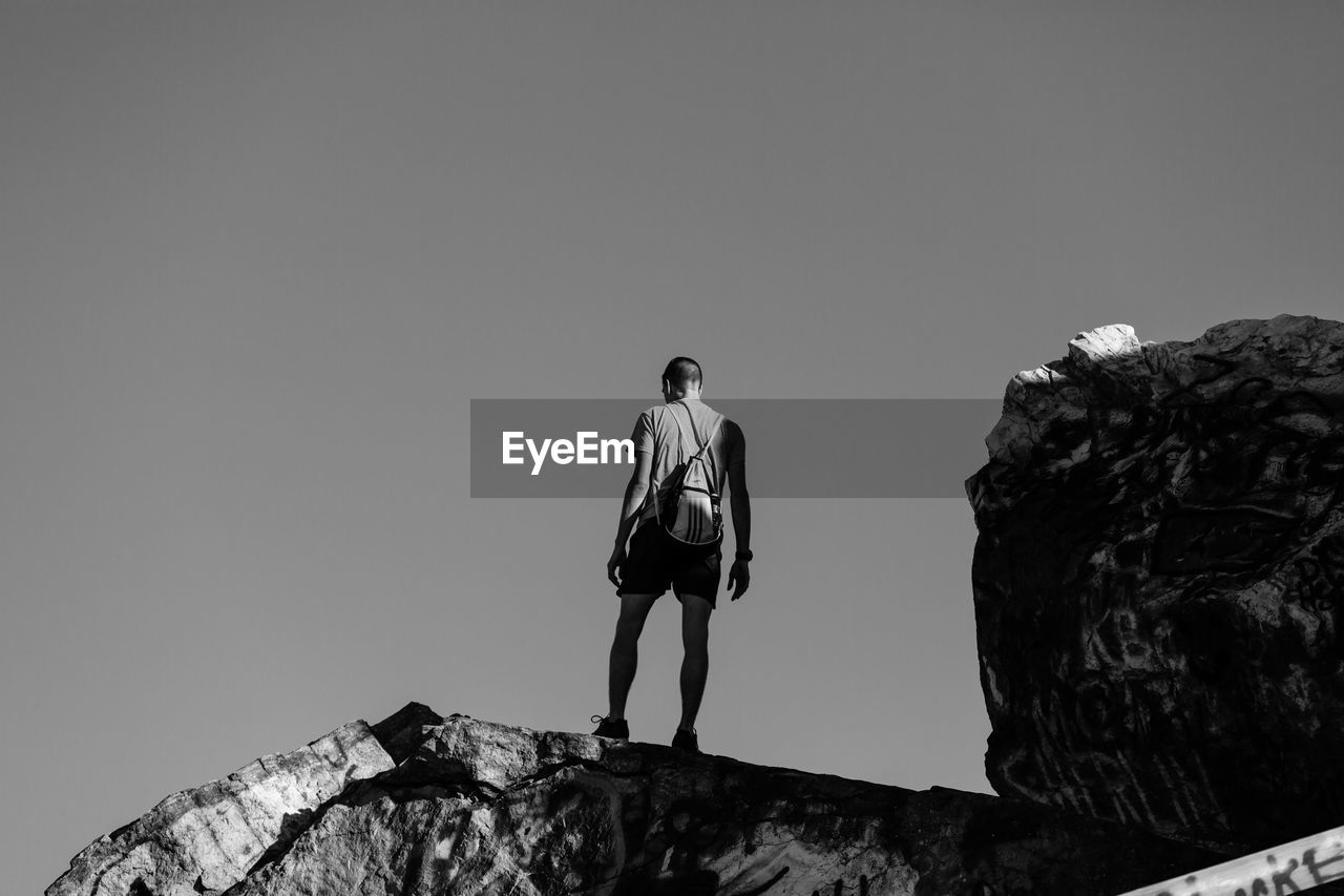 REAR VIEW OF MAN STANDING ON ROCK AGAINST SKY