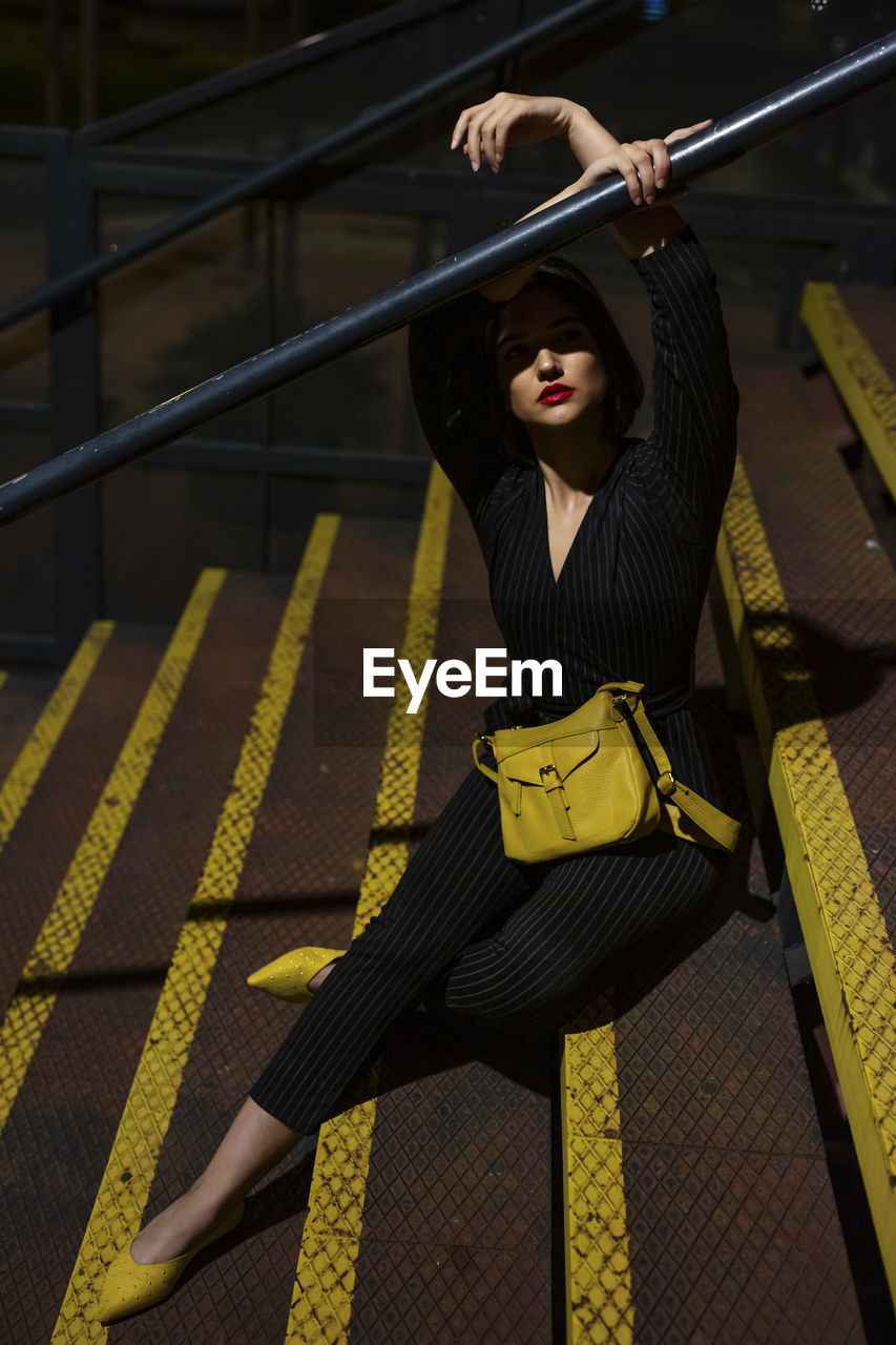 Fashionable woman in black dress with red lipstick and yellow small bag getting into peaky cap sitting at staircase handrail in a city street on dusk