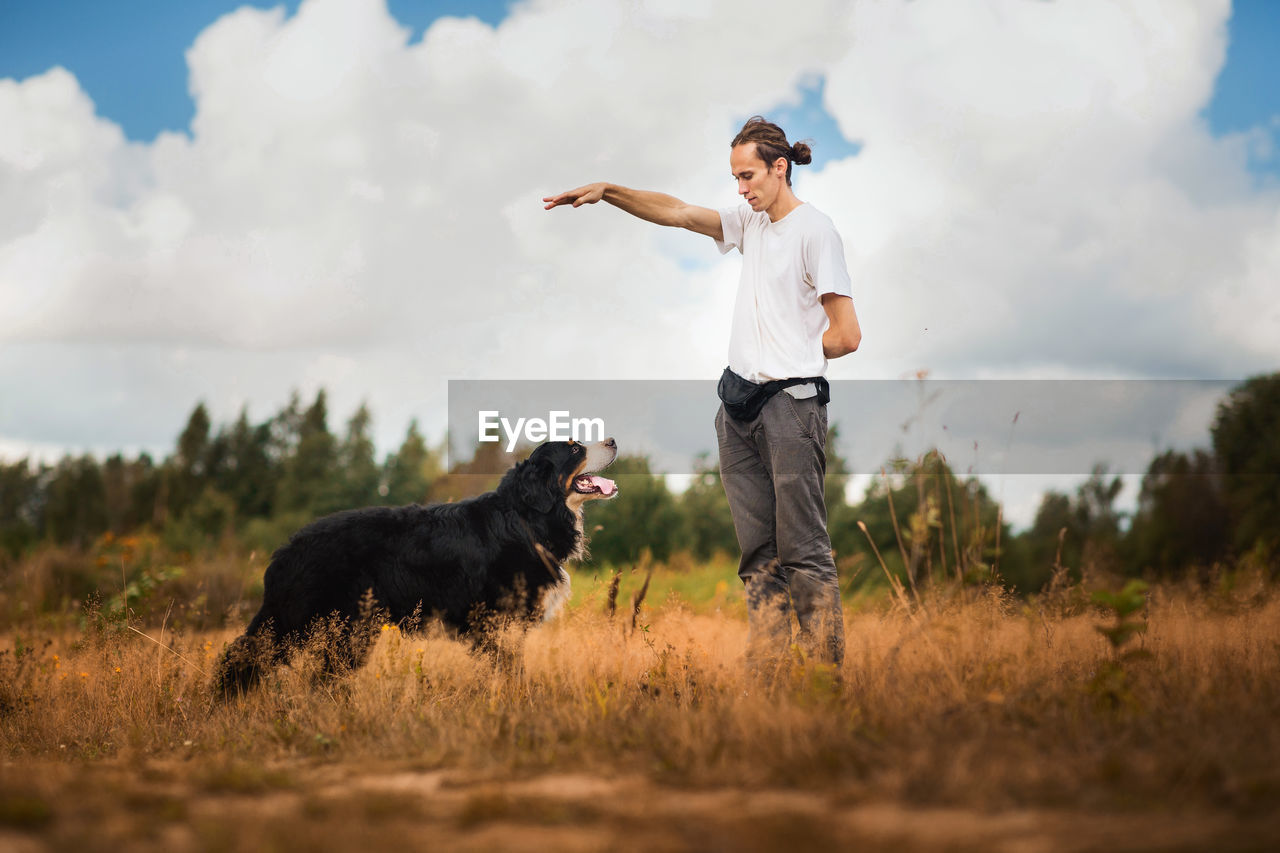 Man training dog on grassy land against cloudy sky