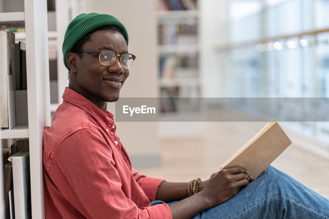 Black student man library visitor sitting on floor near bookcase with book in hands, looks at camera