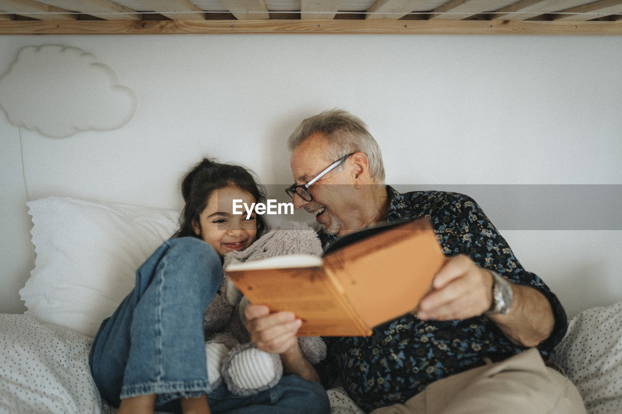 Happy senior man reading storybook with granddaughter on bed at home