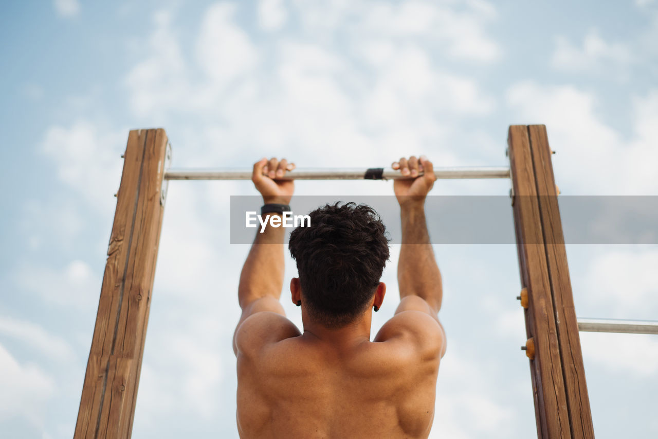 Low angle back view of unrecognizable muscular hispanic ethnic male athlete with naked torso doing chin ups on horizontal bar during training on background of clear blue sky