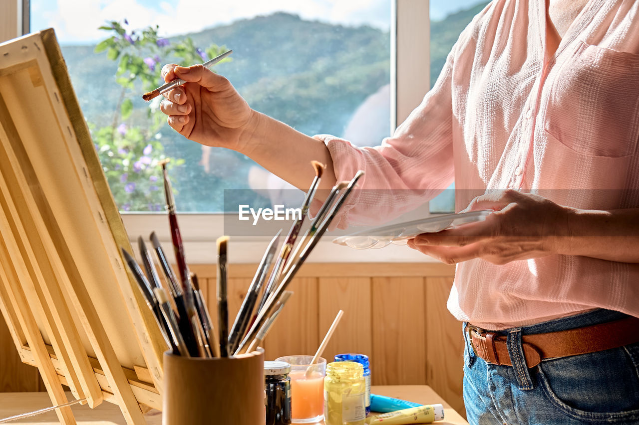 Female hand holding a paintbrush near canvas on easel near the window in the studio.