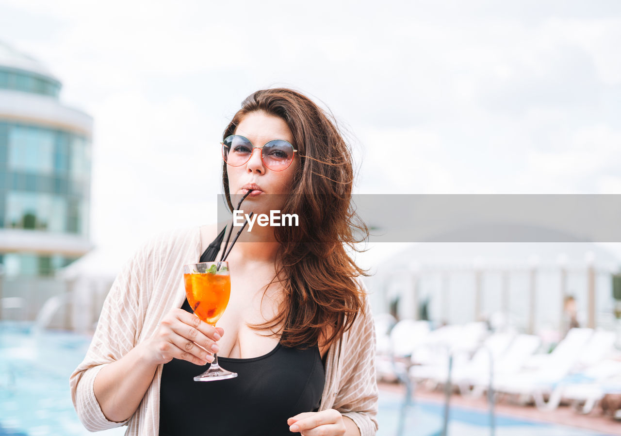 Stylish happy young woman in black swimsuit and sunglasses drinking cocktail near pool