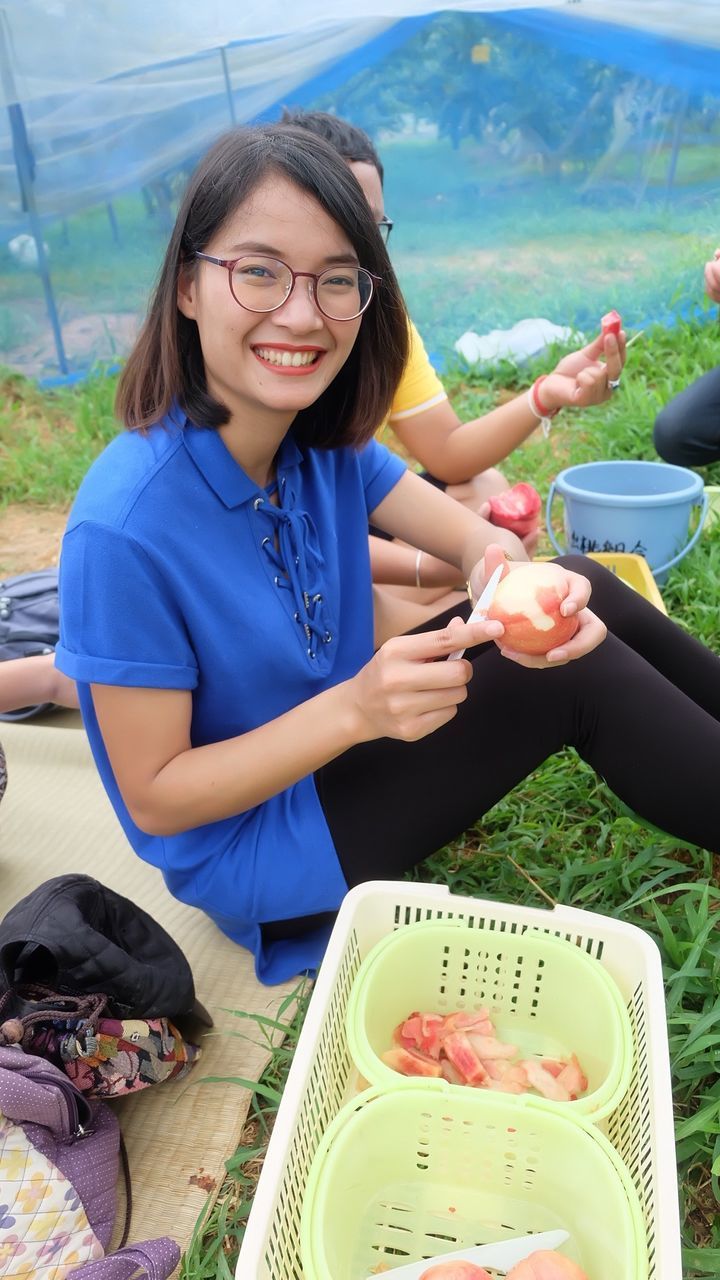 Portrait of smiling young woman peeling apple while sitting on field