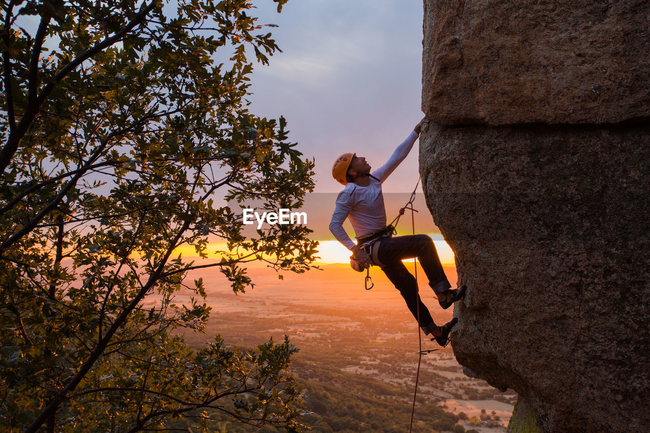 Side view of active climber in protective helmet hanging on mountain holding on to slope in warm light of sunset