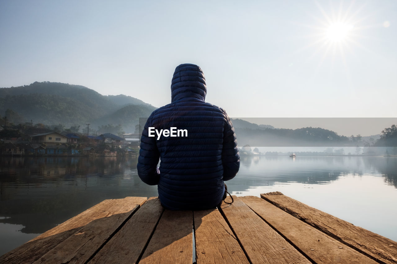 Rear view of man sitting on pier over lake against sky