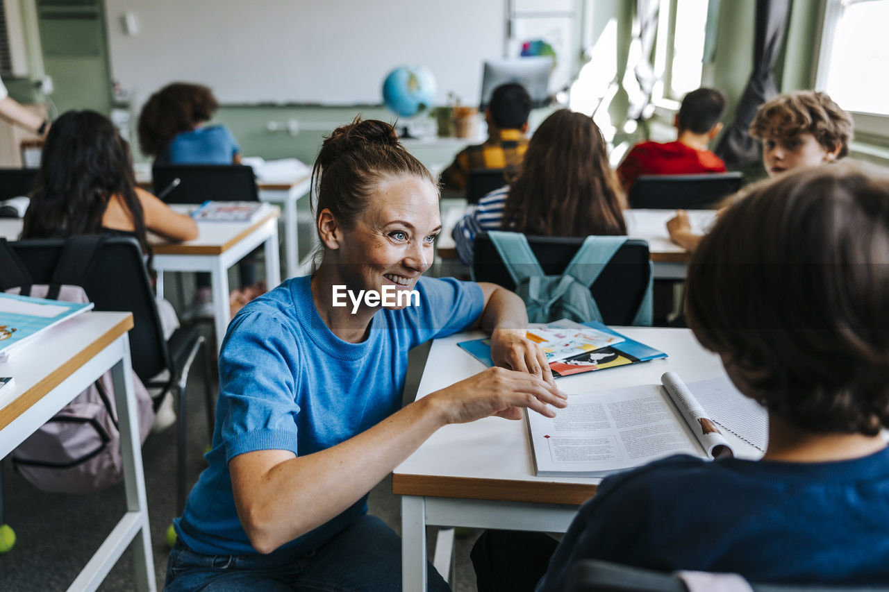 Smiling female teacher assisting student while crouching near desk in classroom at school