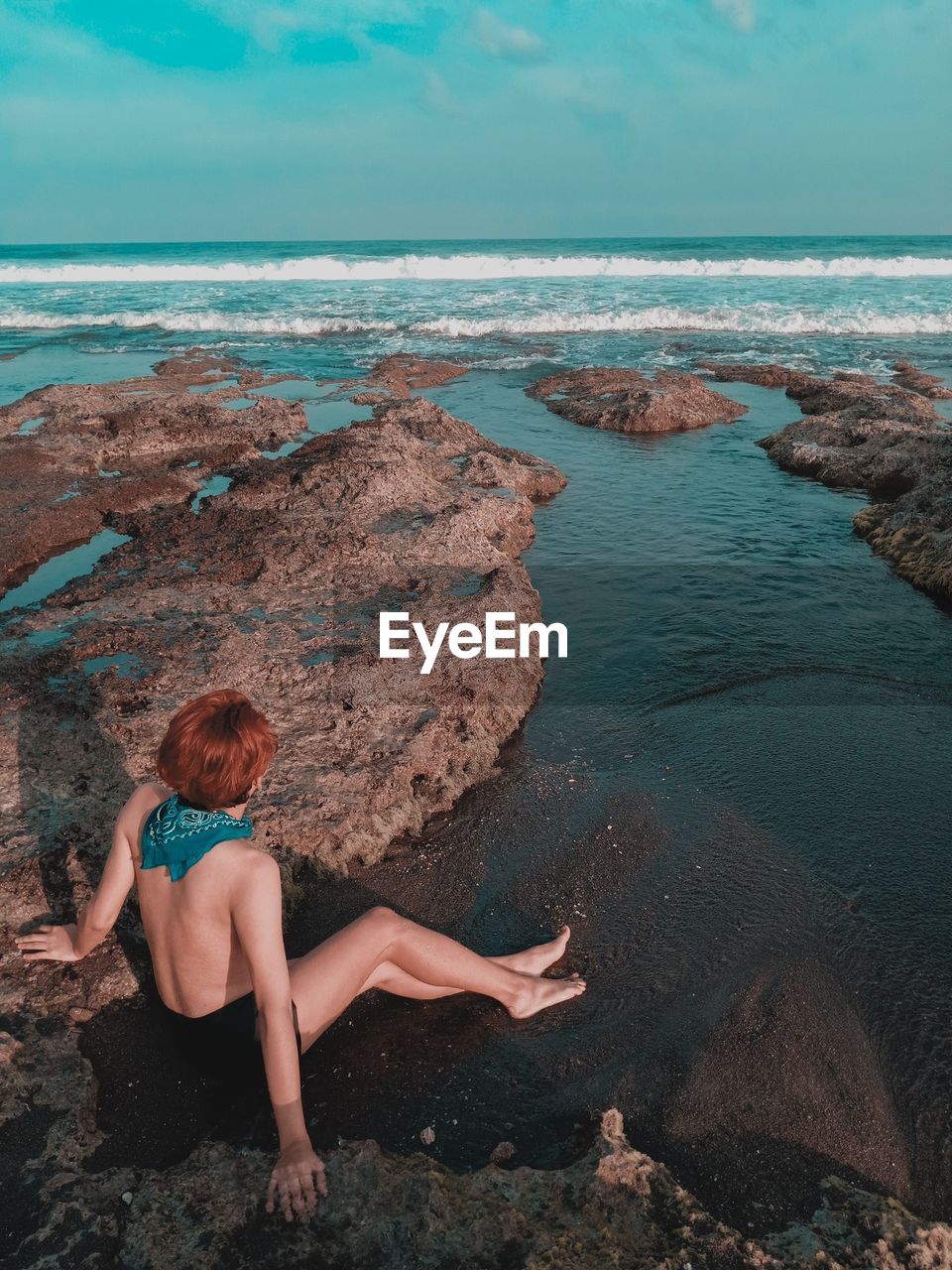 Full length of shirtless man sitting by rock formation at beach