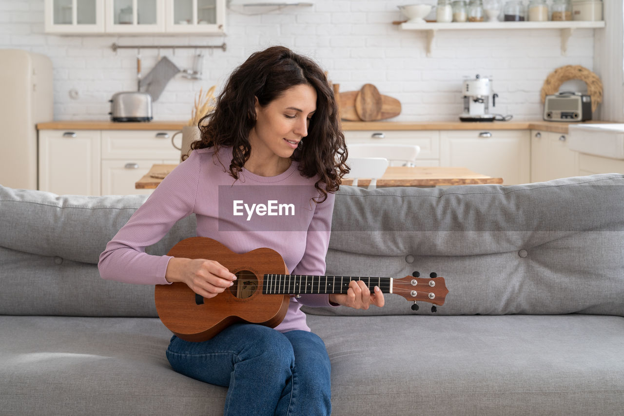 Woman recording content to vlog, playing on ukulele guitar looking at webcam during lockdown at home