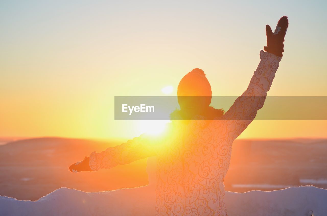 Rear view of woman with hand raised standing against sky during sunset