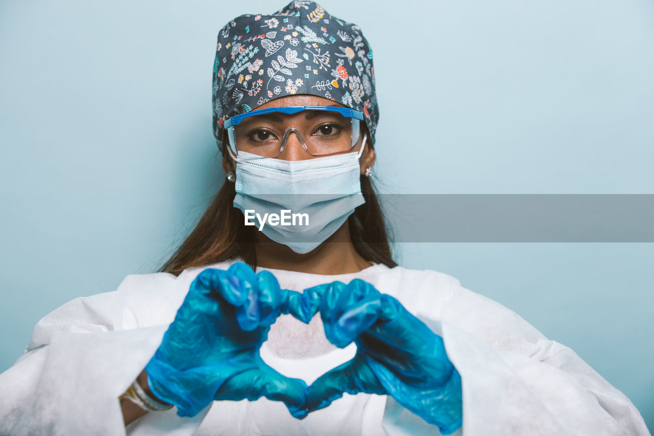 Portrait of young woman wearing mask making heart shape against colored background