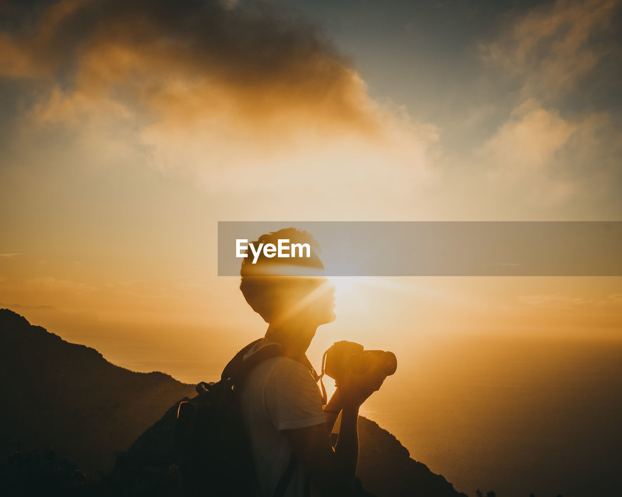  man holding camera standing on mountain against sky during sunset