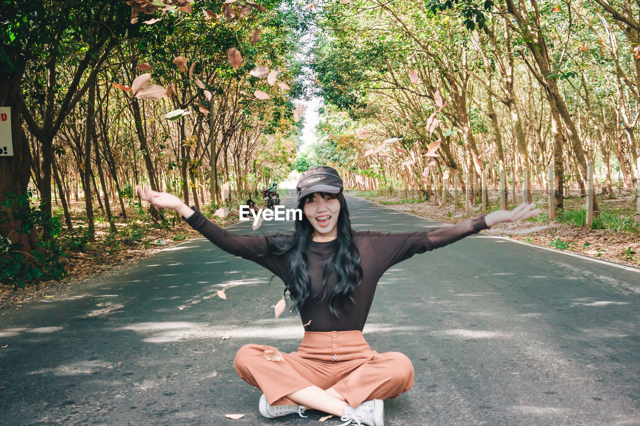 Portrait of smiling young woman throwing leaves while sitting on road amidst trees in forest