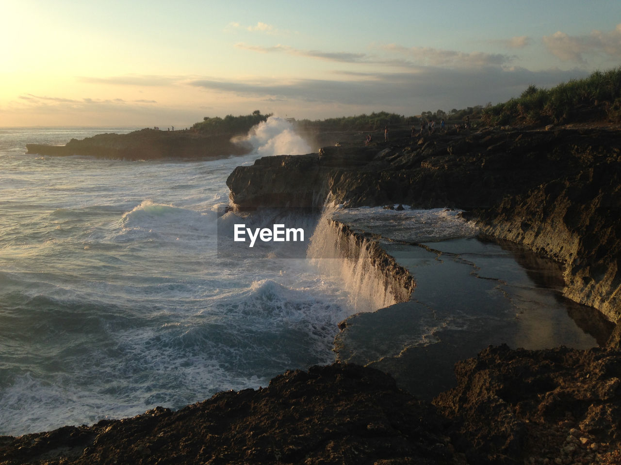 Scenic view of waves splashing on cliff against sky during sunset