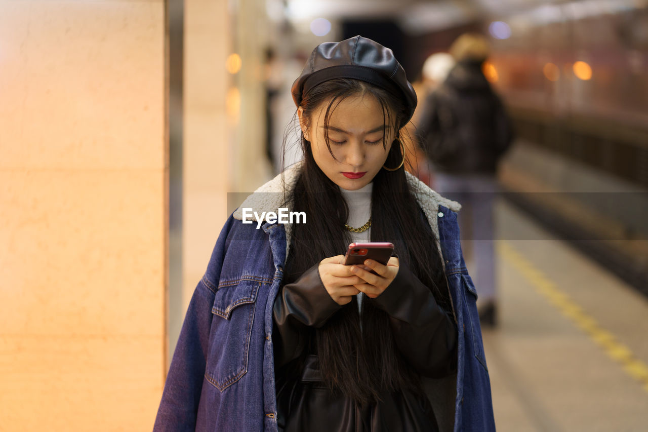 Korean young female texting online at subway metro station hold smartphone, read sms or book tickets