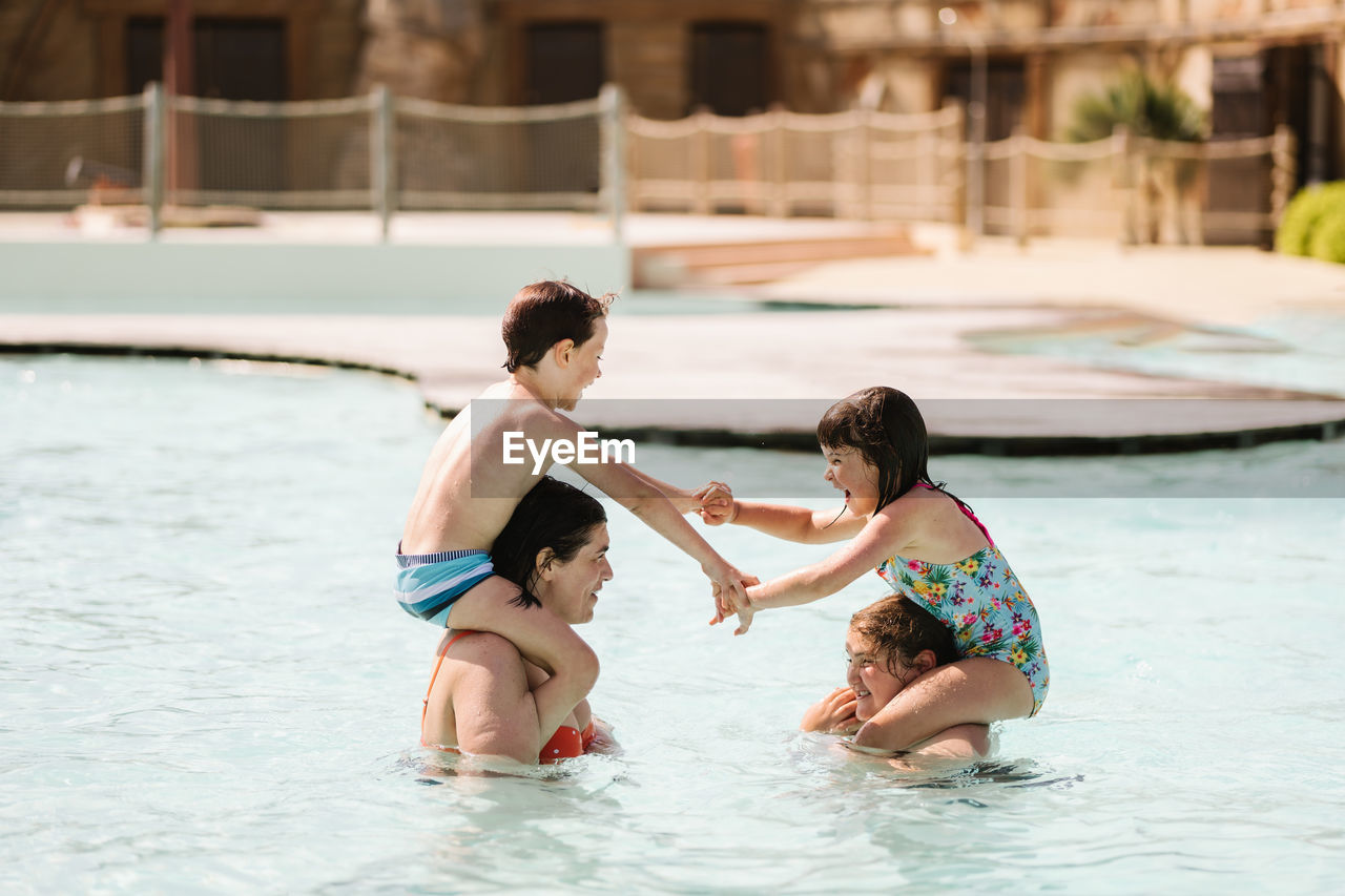 Side view of cheerful little boy and girl in swimwear holding hands and laughing while sitting on shoulders of mother and older sister standing in outdoor swimming pool in sunlight