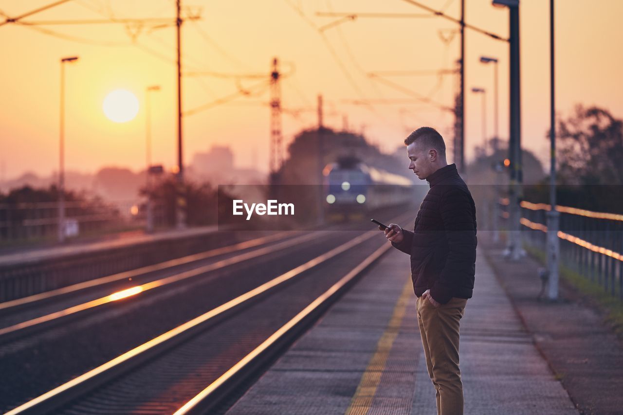 Side view of man standing at railroad station against sky during sunset