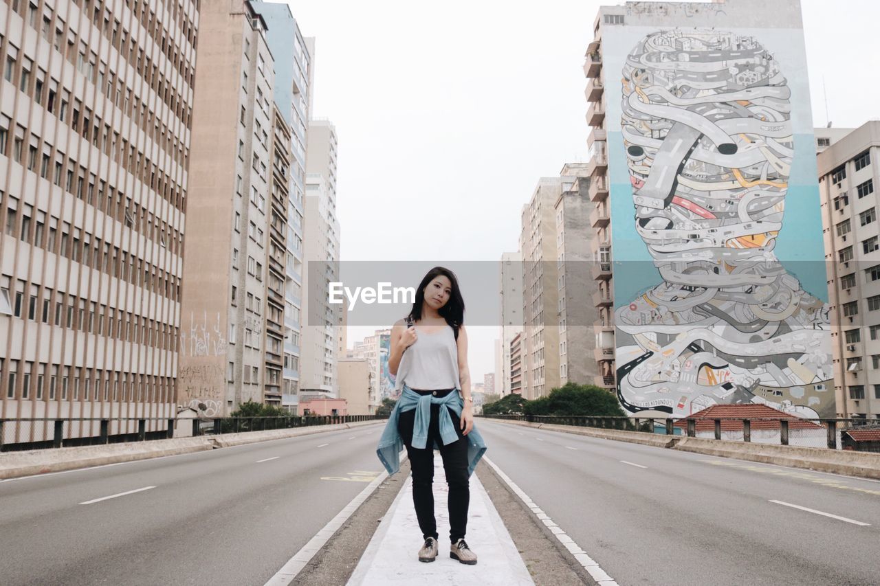 PORTRAIT OF WOMAN STANDING ON ROAD IN CITY