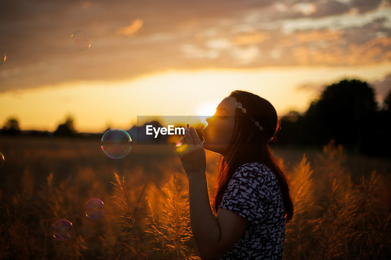 Young woman blowing bubbles on wheat field against cloudy sky during sunset