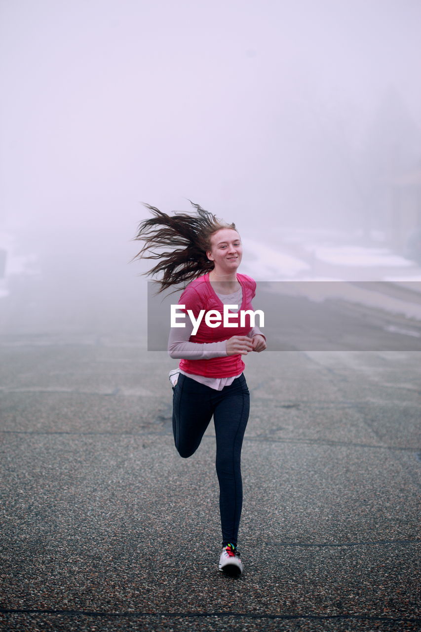 Portrait of smiling young woman running on road during foggy weather