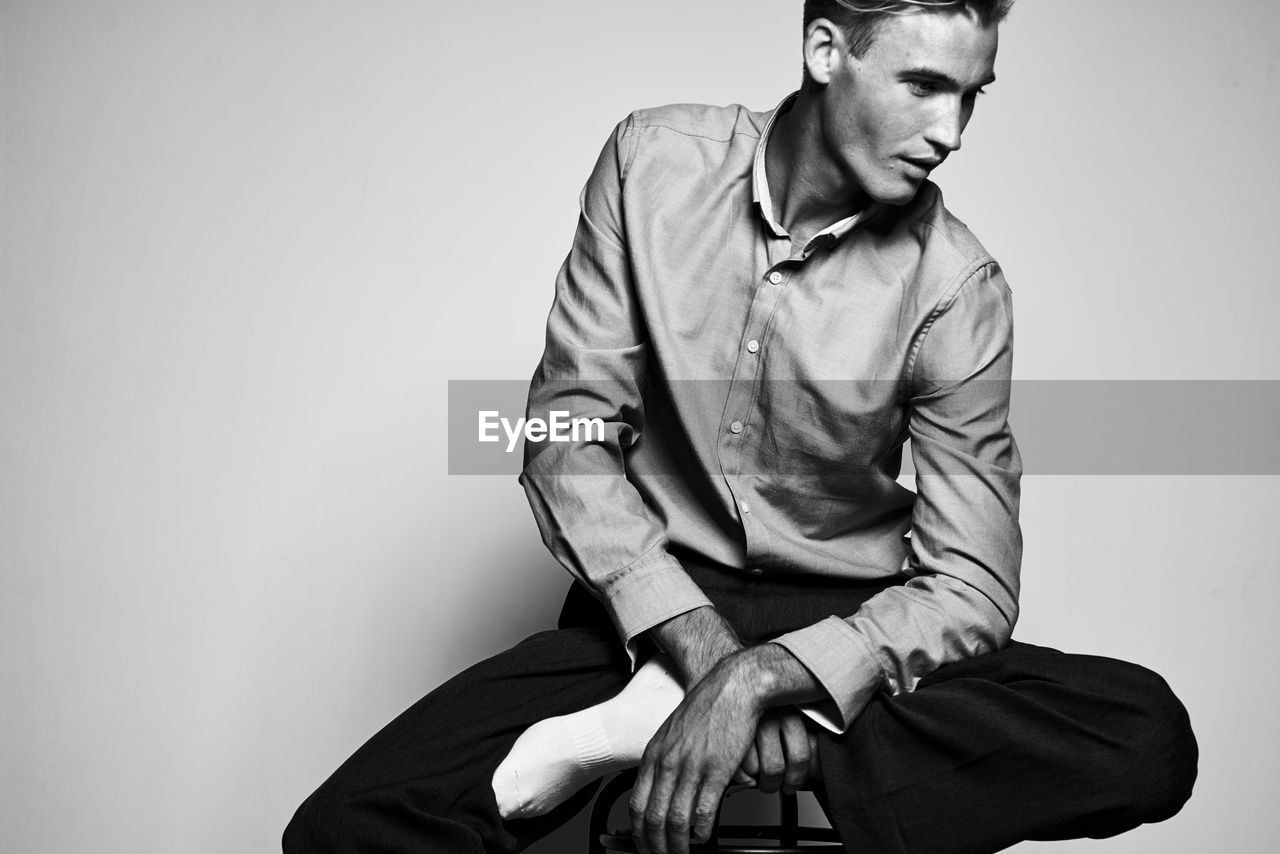 YOUNG MAN LOOKING AWAY WHILE SITTING ON WALL