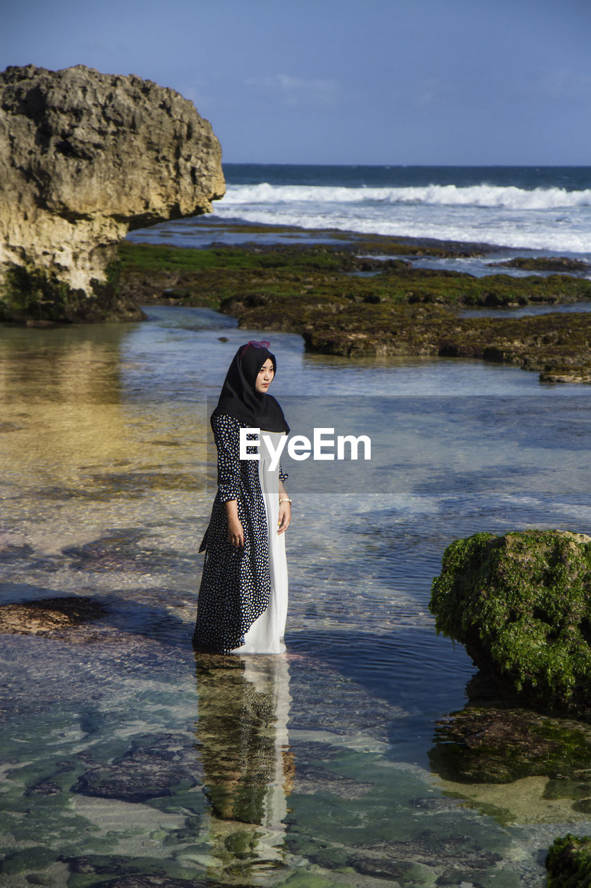 WOMAN STANDING ON ROCK IN SEA