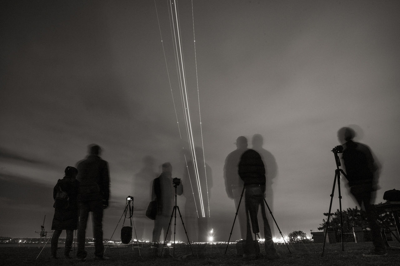 Double exposure of people standing by cameras on tripod against sky
