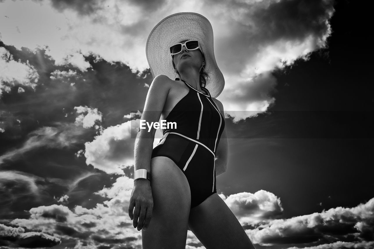 A portrait of a woman in a swimsuit, hat and sunglasses in summer on the riverbank against the sky