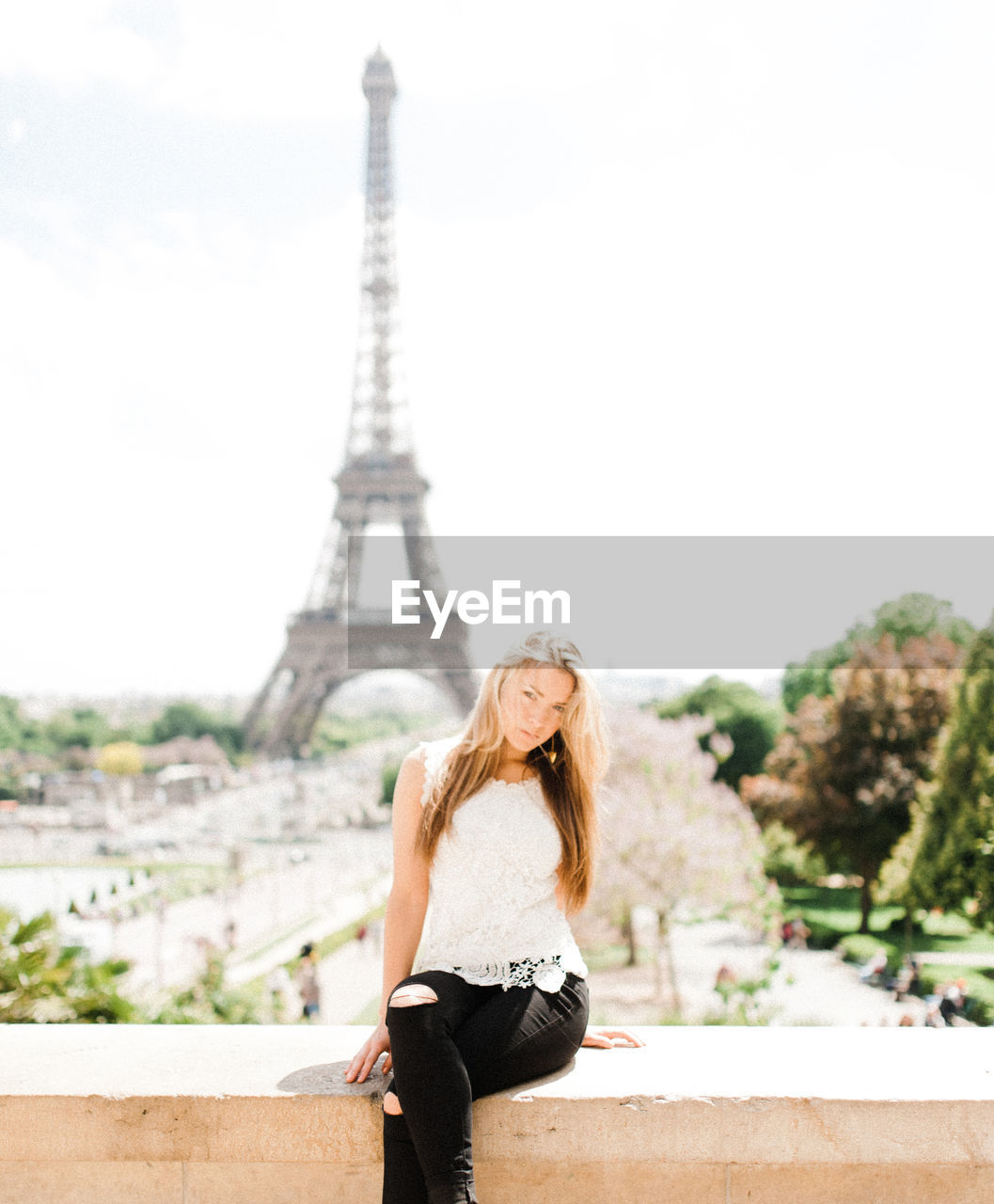 Portrait of young woman sitting on concrete wall with eiffel tower in background