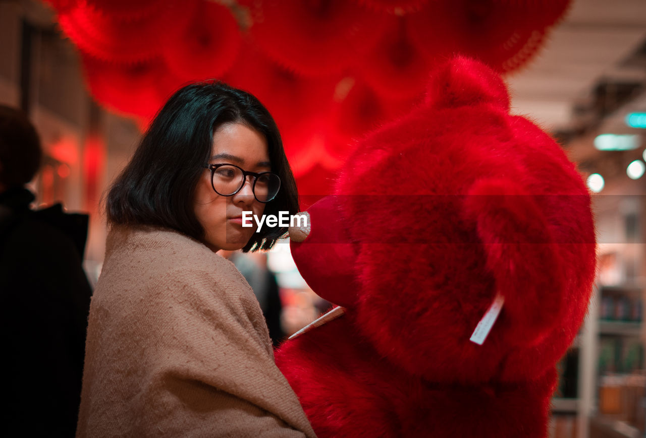 Young woman holding red teddy bear while standing in room