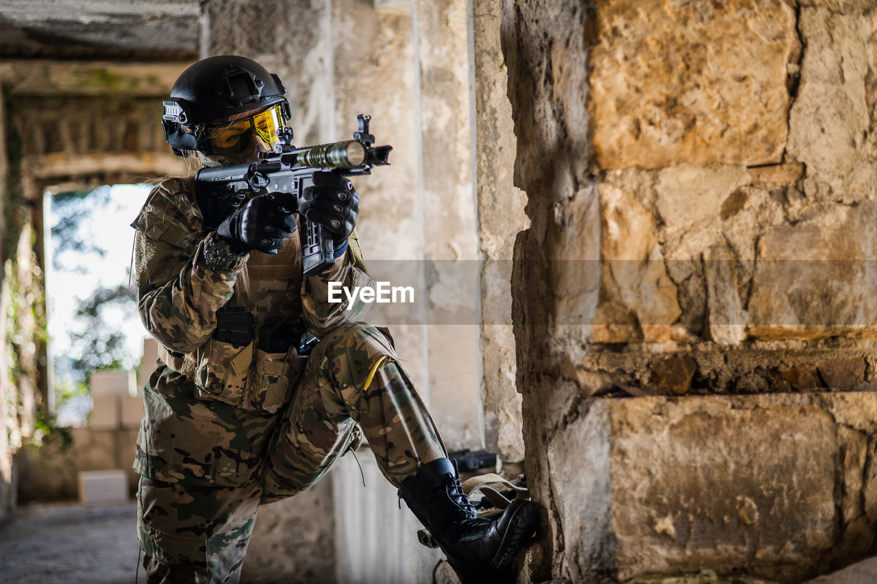 A woman in an army uniform aims to shoot a firearm in an abandoned building. 