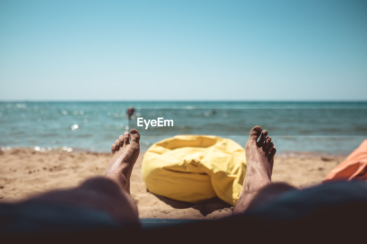 Low section of man relaxing on beach against clear sky