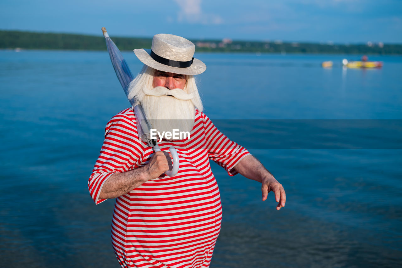 MIDSECTION OF WOMAN WEARING HAT STANDING BY WATER