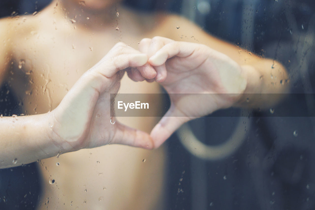 Midsection of shirtless boy making heart shape seen through wet bathroom glass