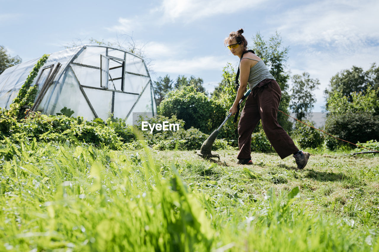 Woman cutting grass with a trimmer and a greenhouse in. the background