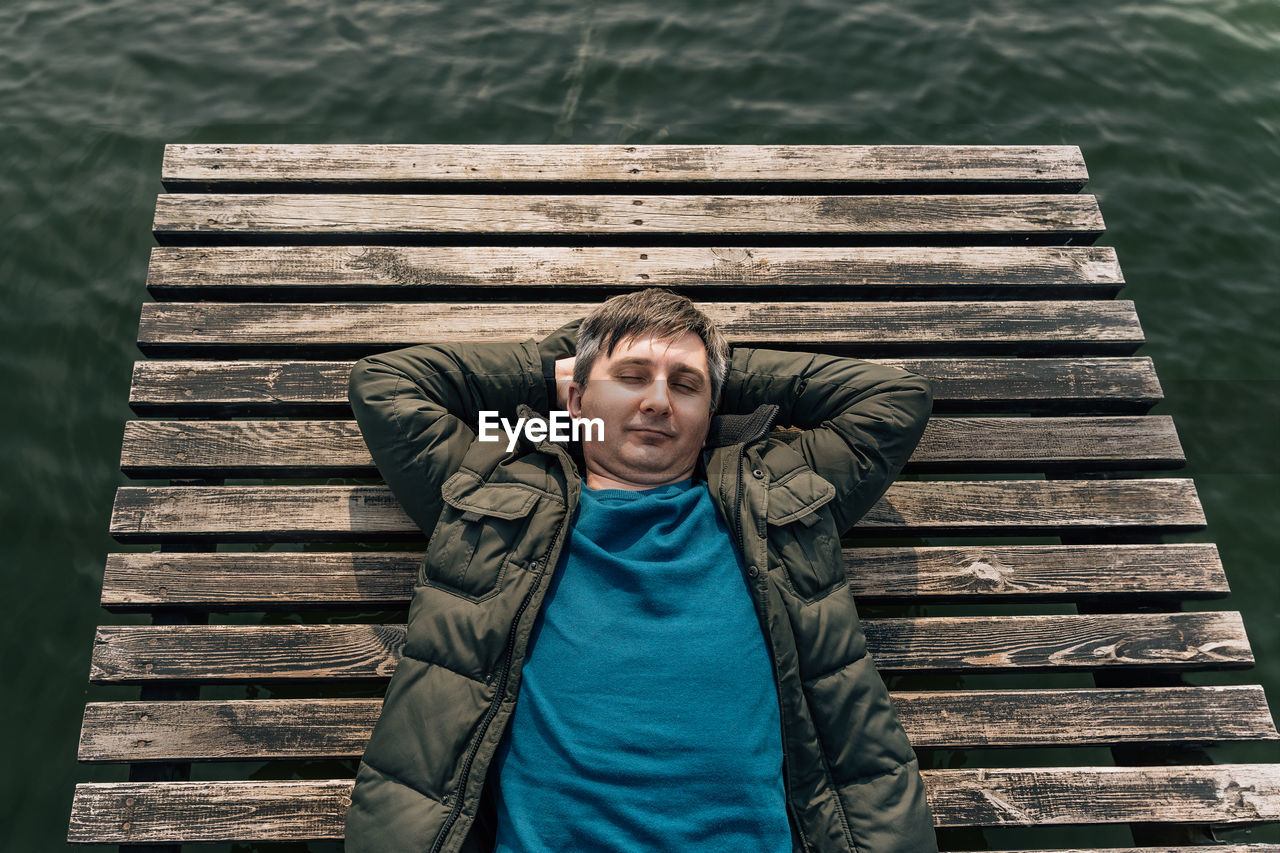 A man lies on a wooden plank pier on the lake in a jacket and jumper with his eyes closed