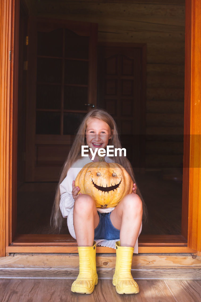 A cheerful girl with long hair is sitting on a doorstep, holding a pumpkin with eyes and mouth 