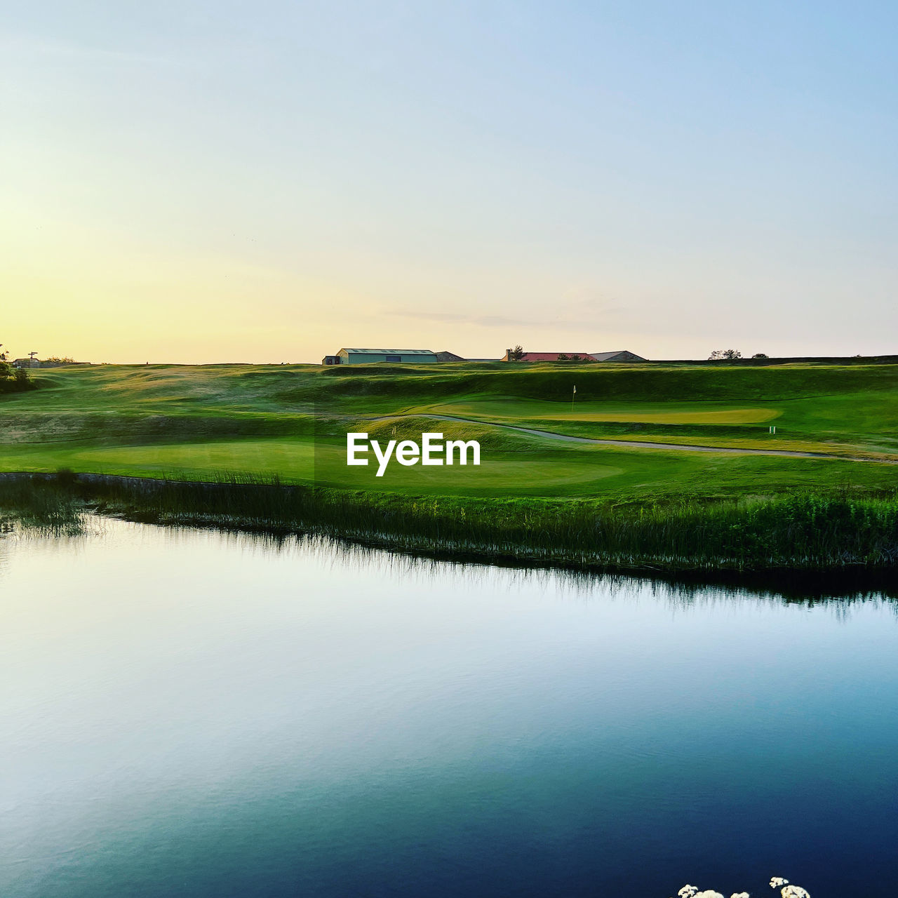 water, sky, reflection, scenics - nature, green, grass, environment, nature, landscape, lake, golf, beauty in nature, tranquility, plant, tranquil scene, grassland, no people, activity, sport venue, golf course, sports, clear sky, land, non-urban scene, leisure activity, plain, copy space, outdoors, horizon, cloud, field, rural scene, day, golf club, blue, idyllic, sunset, tree