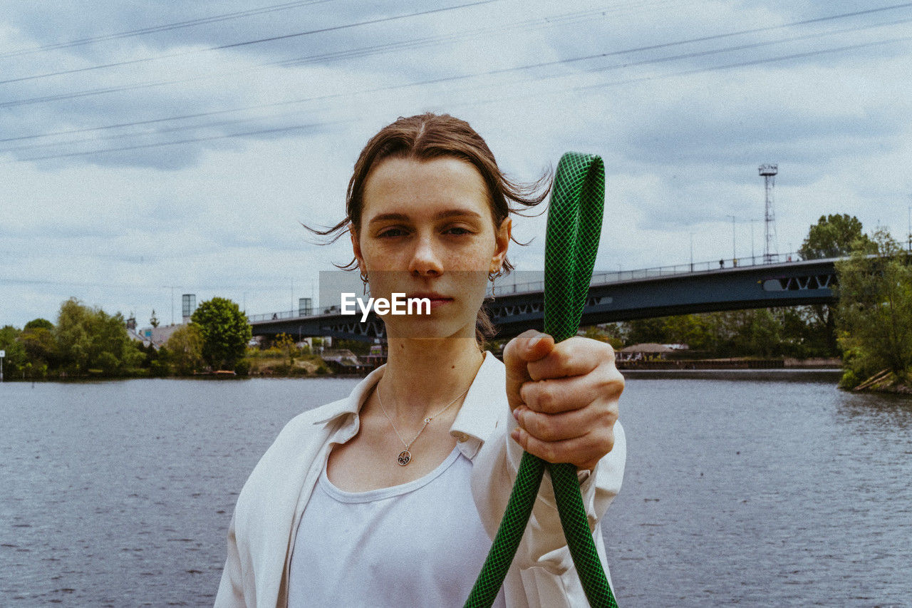 Portrait of confident young non-binary person holding garden hose near river
