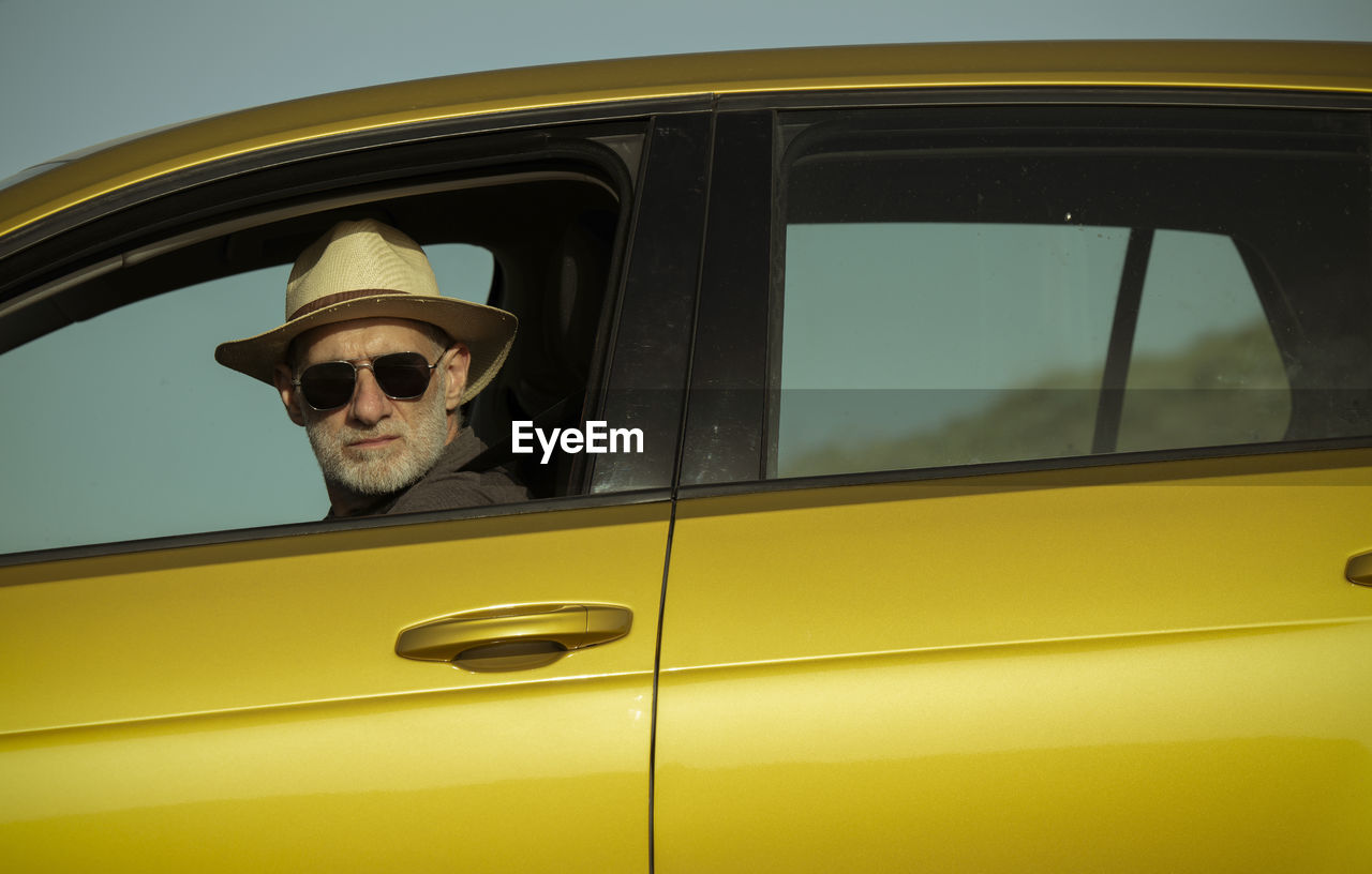 Adult man in hat and sunglasses sitting in car against clear sky