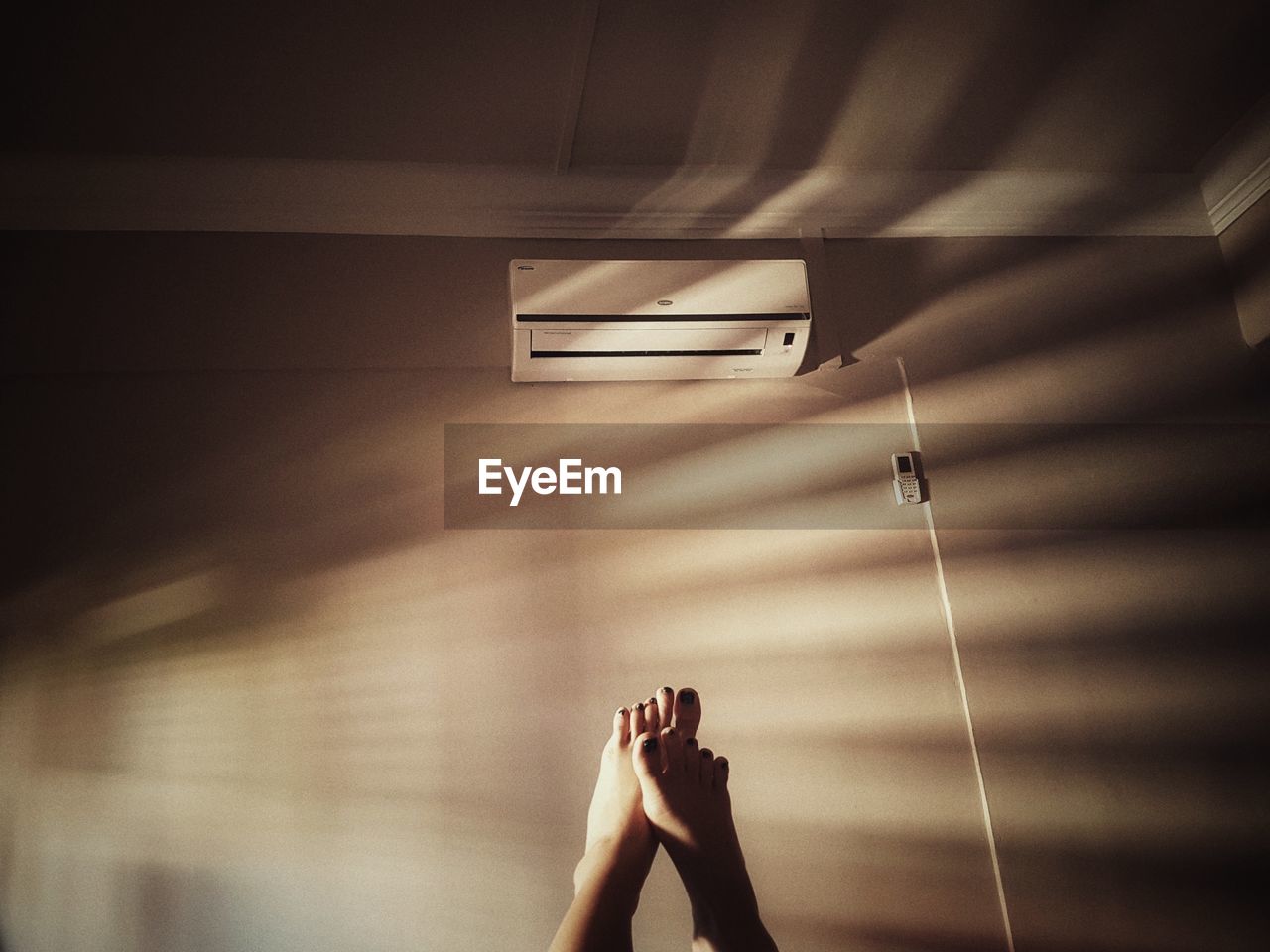 Low section of woman with feet up against air conditioner on wall at home