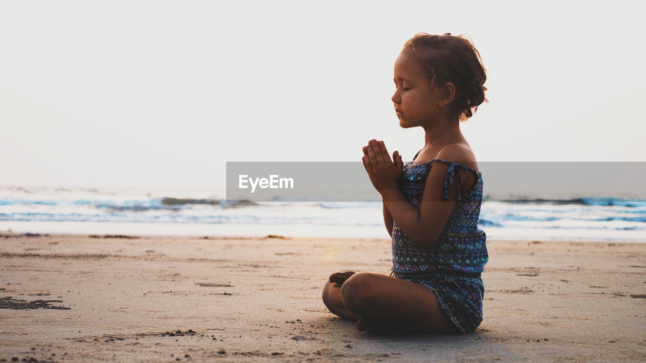 Full length side view of girl praying while sitting on beach against clear sky