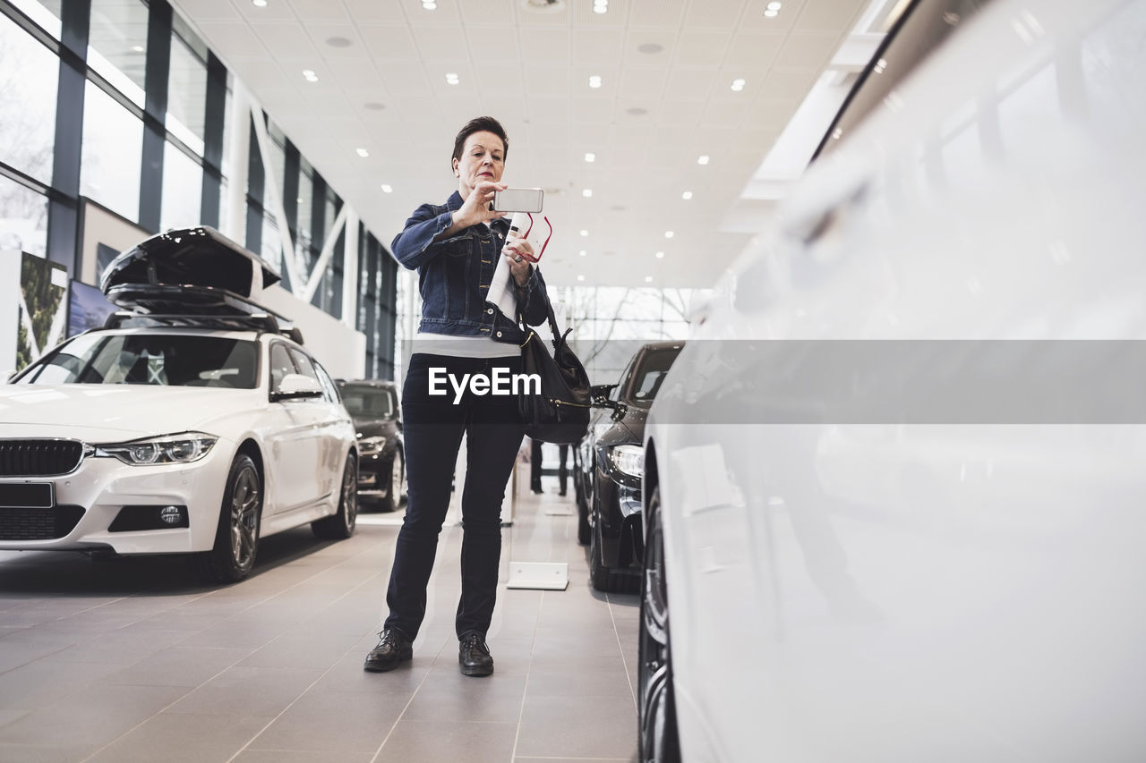 Senior woman photographing new car through mobile phone at showroom