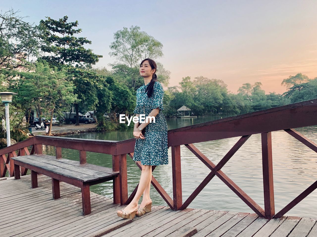 Full length of young woman standing on footbridge over river against sky in park during sunset