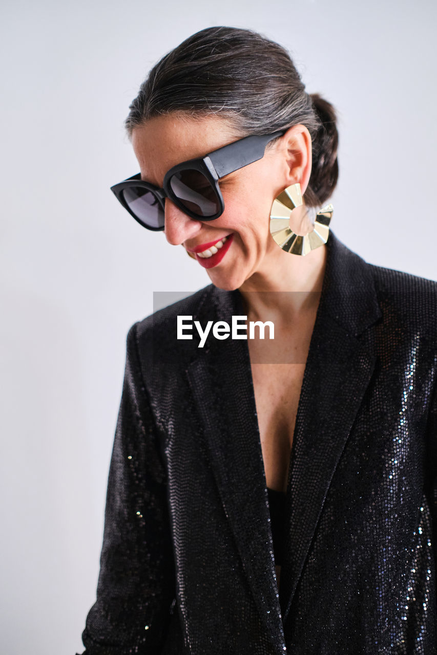 Positive female in stylish black outfit and trendy sunglasses with round earrings standing on white background in modern light studio