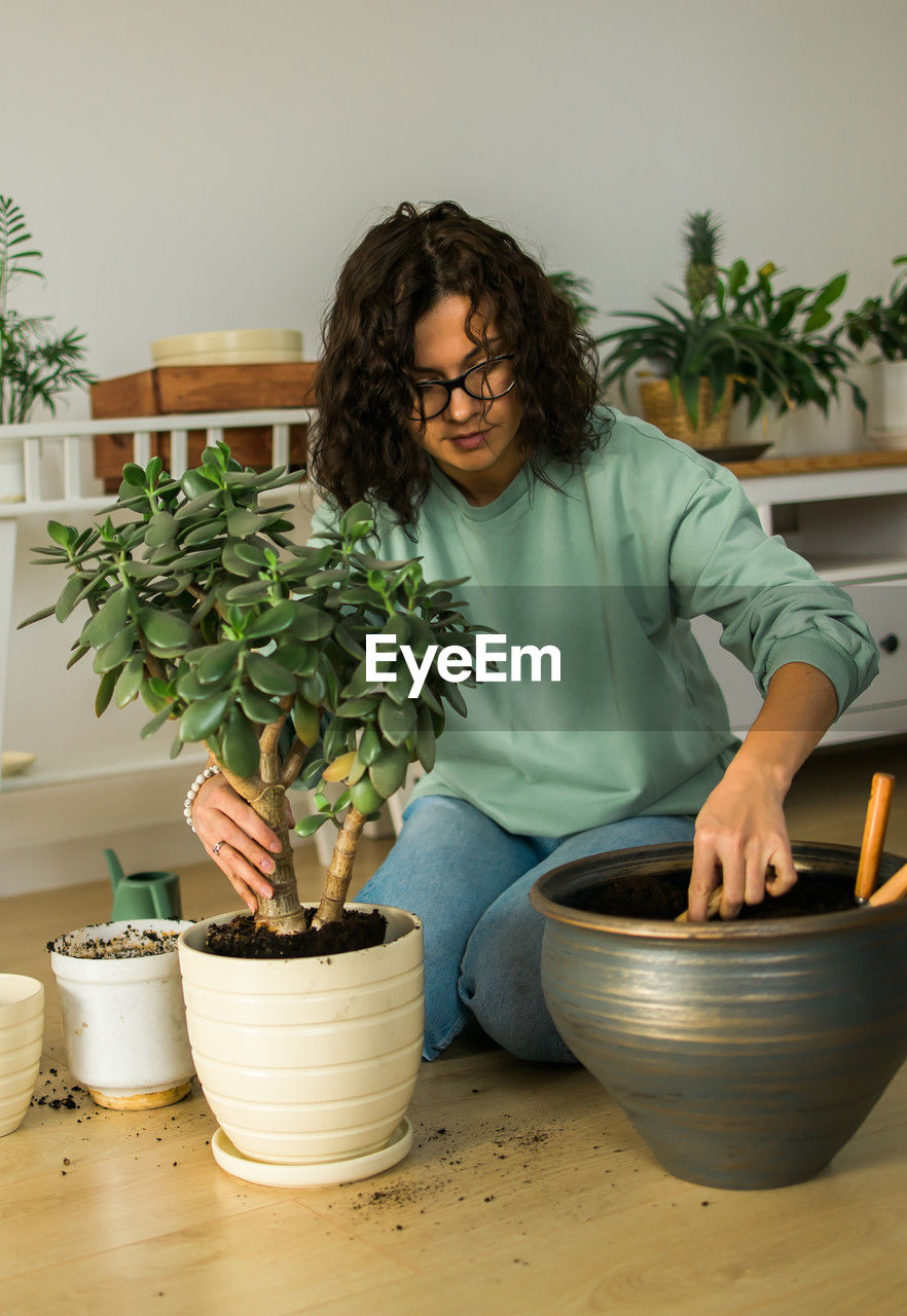 one person, adult, potted plant, indoors, plant, lifestyles, flowerpot, women, front view, domestic life, young adult, growth, nature, hairstyle, brown hair, houseplant, holding, curly hair, casual clothing, domestic room, looking, home interior, sitting, looking down, female, leisure activity, person